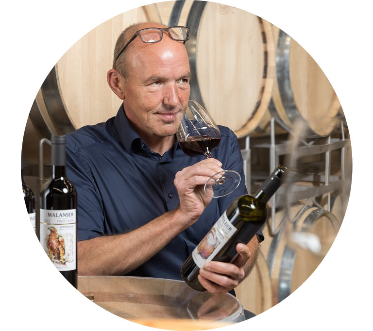 Konzentriert überprüft ein Mitarbeiter den Geruch eines Glases Rotwein. Im Hintergrund sind Holzfässer zu erkennen.